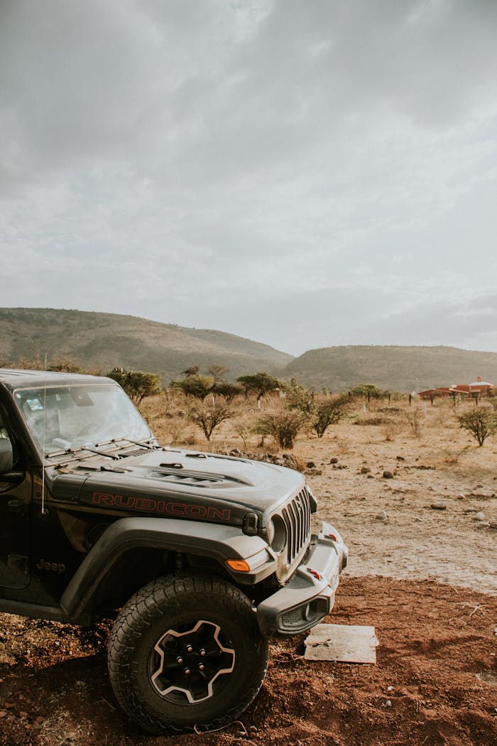A rugged black Jeep Wrangler parked offroad with a scenic field and hills in the background.